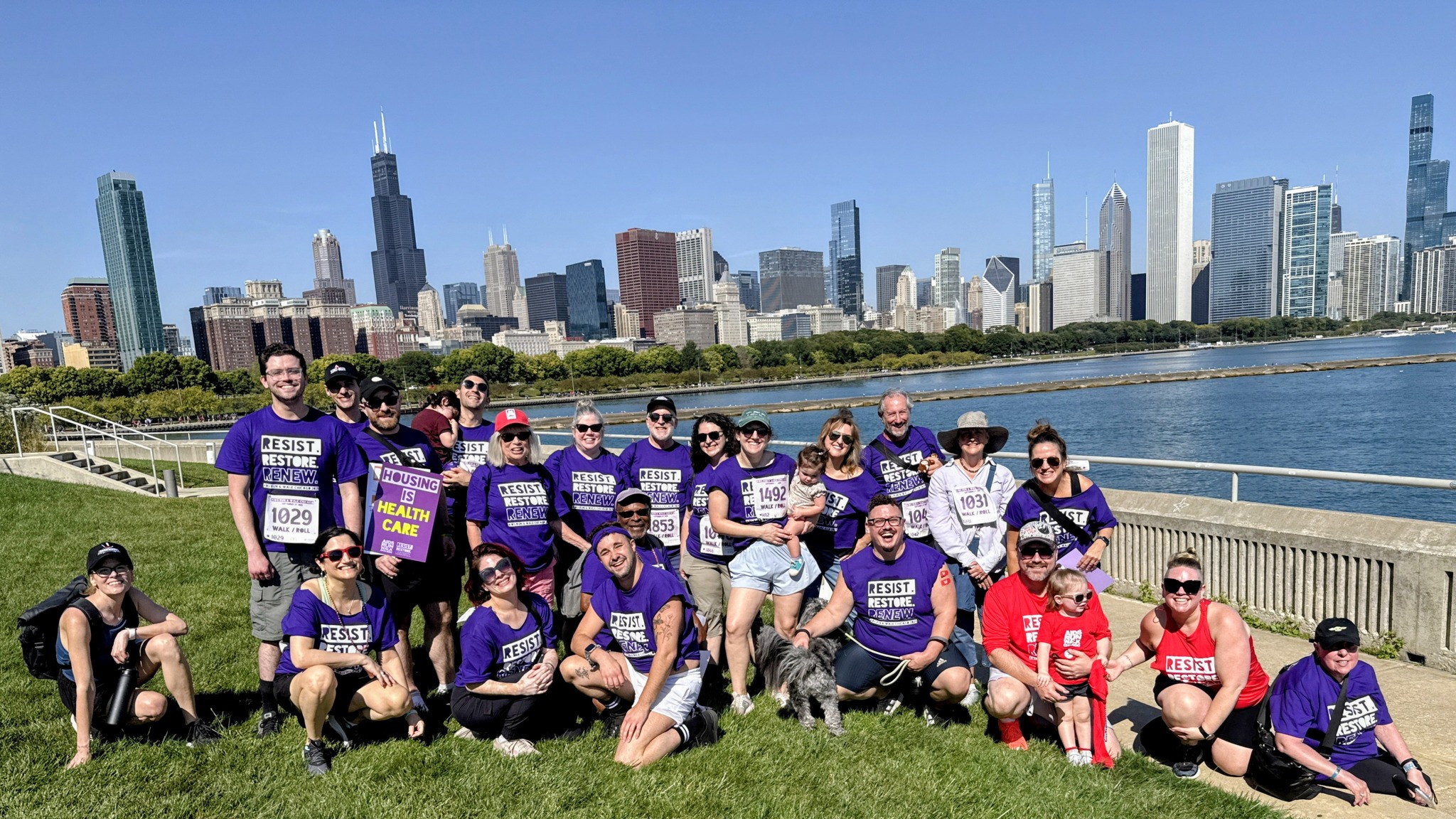 A crowd of about a couple dozen people in shirts that say "RESIST RESTORE RENEW" pose in front of the Chicago waterfront.