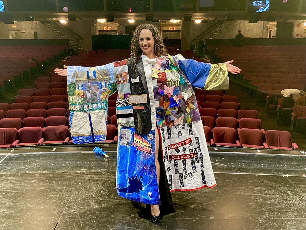 Nina Lafarga poses onstage in the Robe, arms outstretched and smiling, with the house visible behind her.