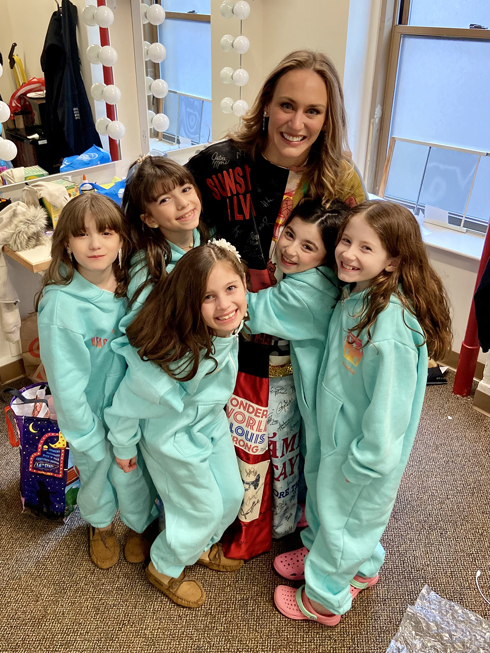 In a dressing room, Martignetti wears the Robe and embraces five young girls, all of whom are wearing matching jumpsuits.