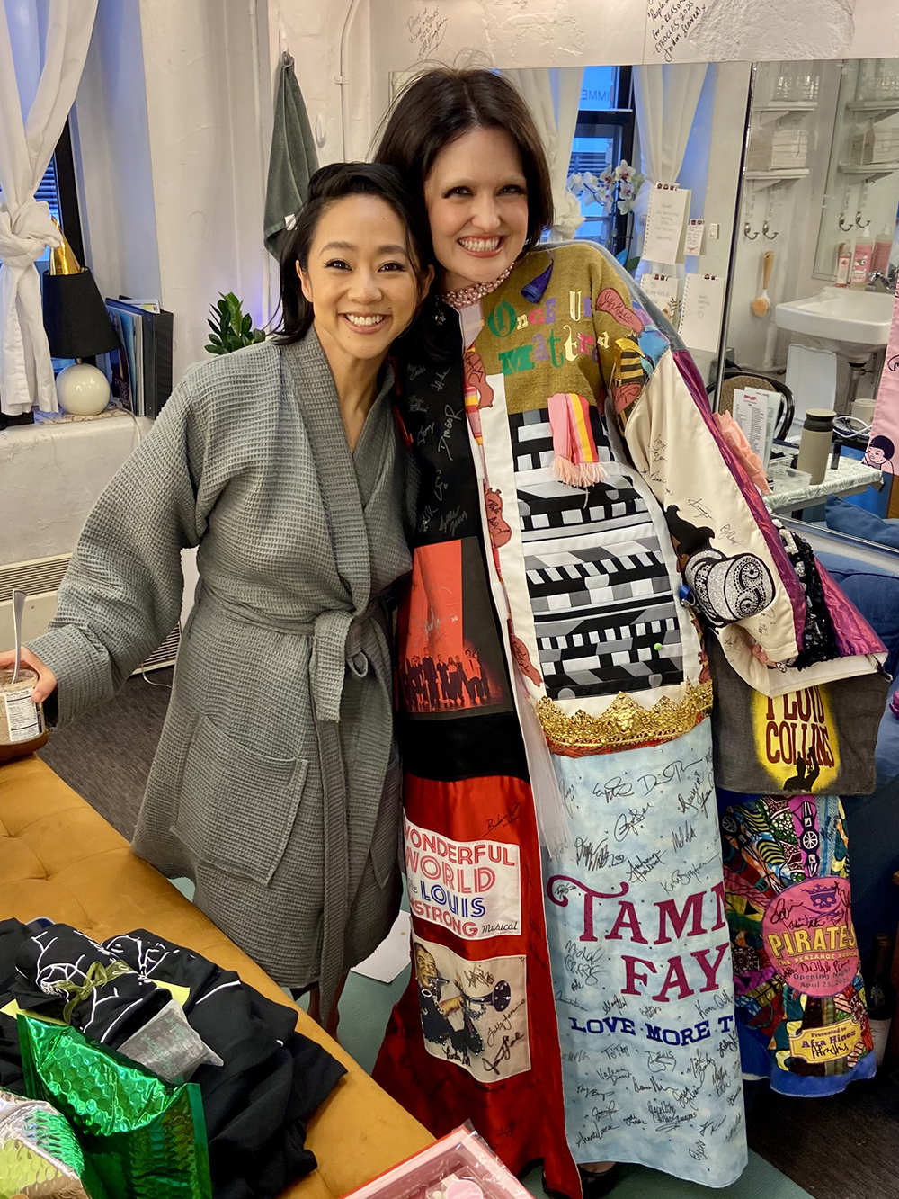 Stephanie Hsu poses in a bathrobe in a dressing room next to Reilly in the Robe. Both are smiling.