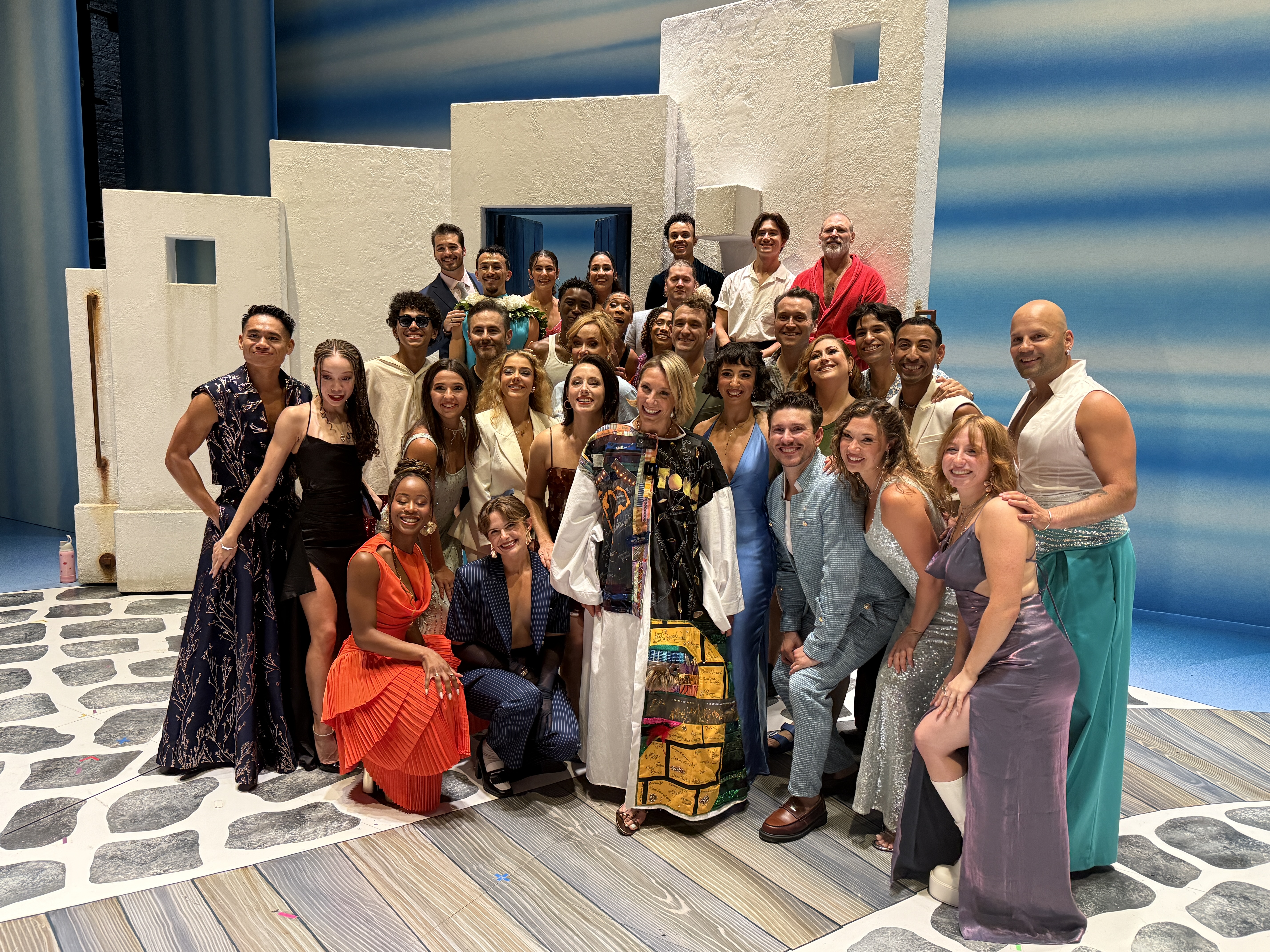 Erica Mansfield stands onstage in the Robe posing with about 30 other people surrounding her. The Mediterranean backdrop is visible behind them