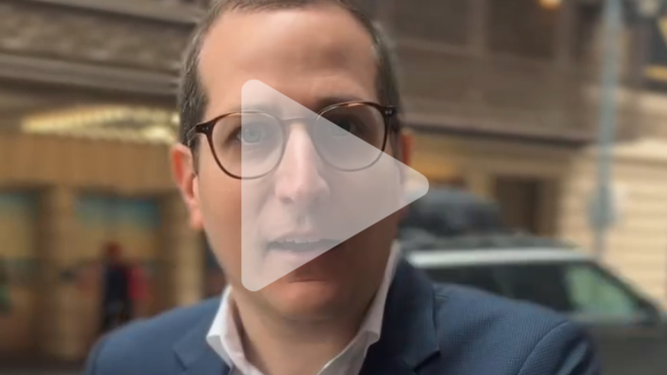 A semi-transparent play button logo over an image of Micah Lasher Speaking