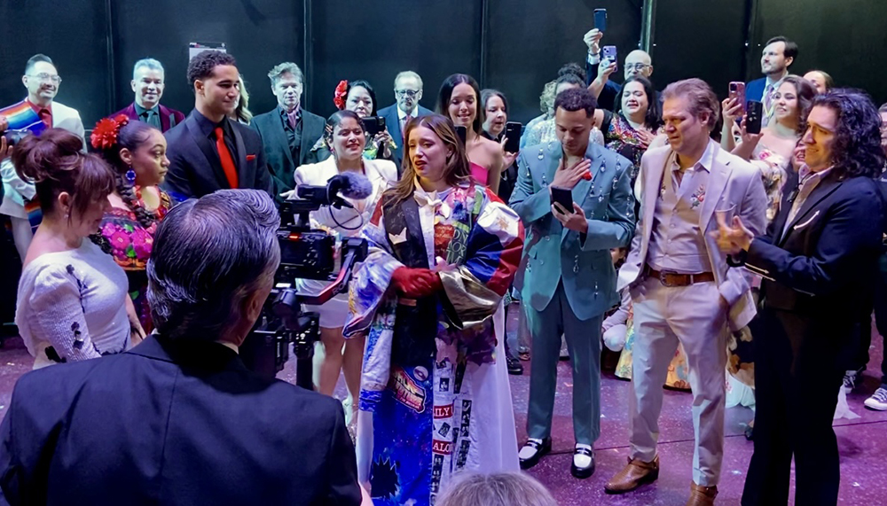 A crowd of people form around Ramirez, who looks emotional in the Robe, facing a visible camera capturing the moment.