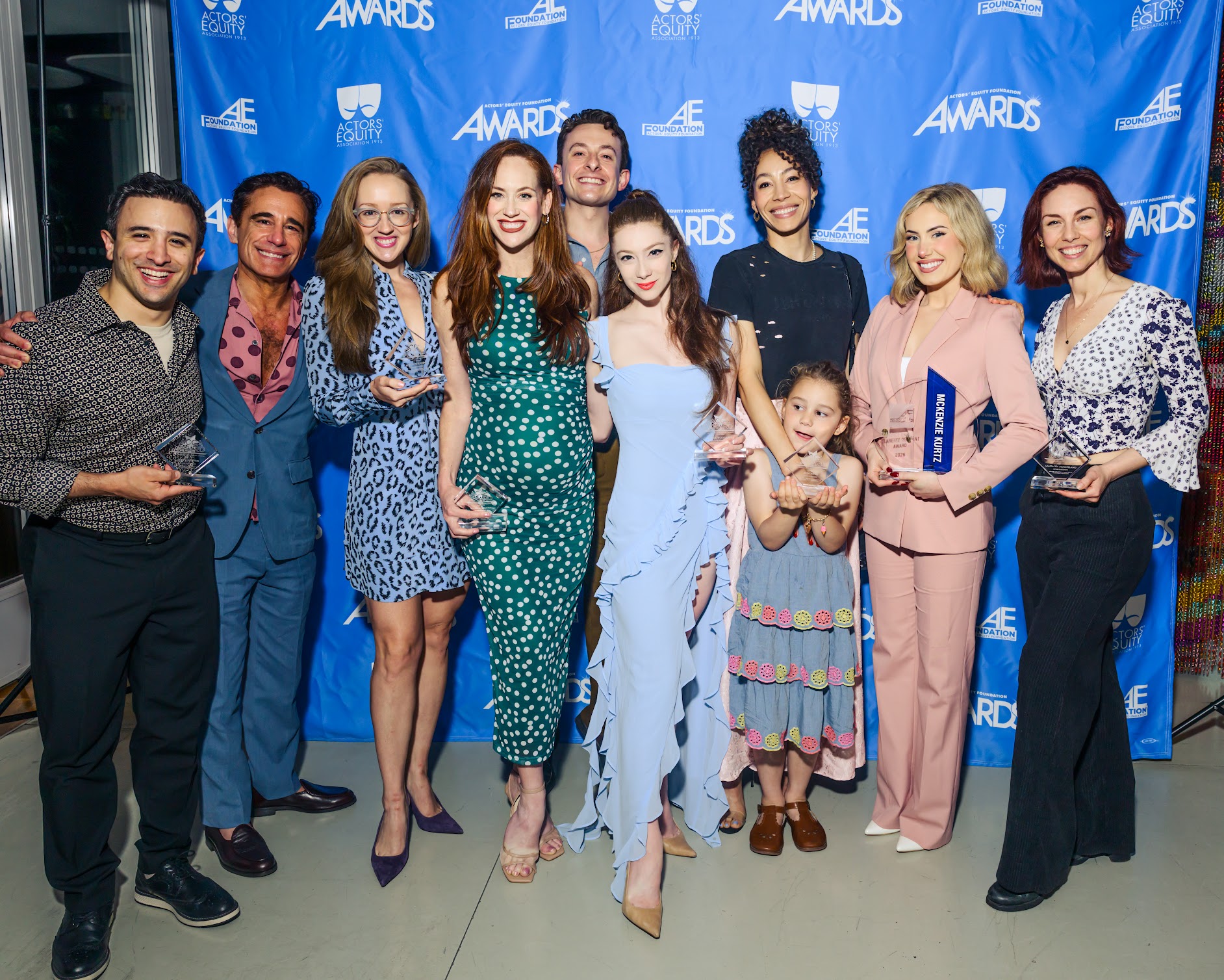 A group of nine adults (many of whom are holding trophies) and one child pose and smile in front of the Actors' Equity Foundation Awards step-and-repeat.