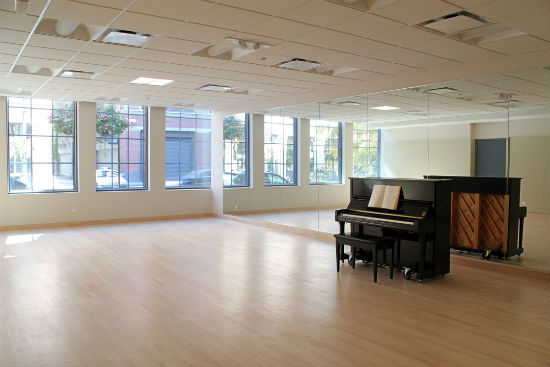 A large room with many light-filled windows, a wall-length mirror, an upright piano and wood floor