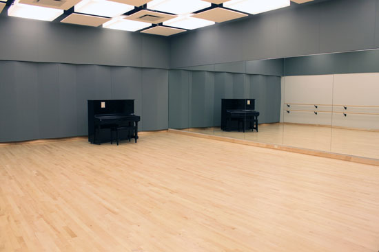 Large Audition Room with soundproofing on the far wall, an upright piano in the corner, a mirror on the near wall and barres visible in the reflection of the opposite wall.